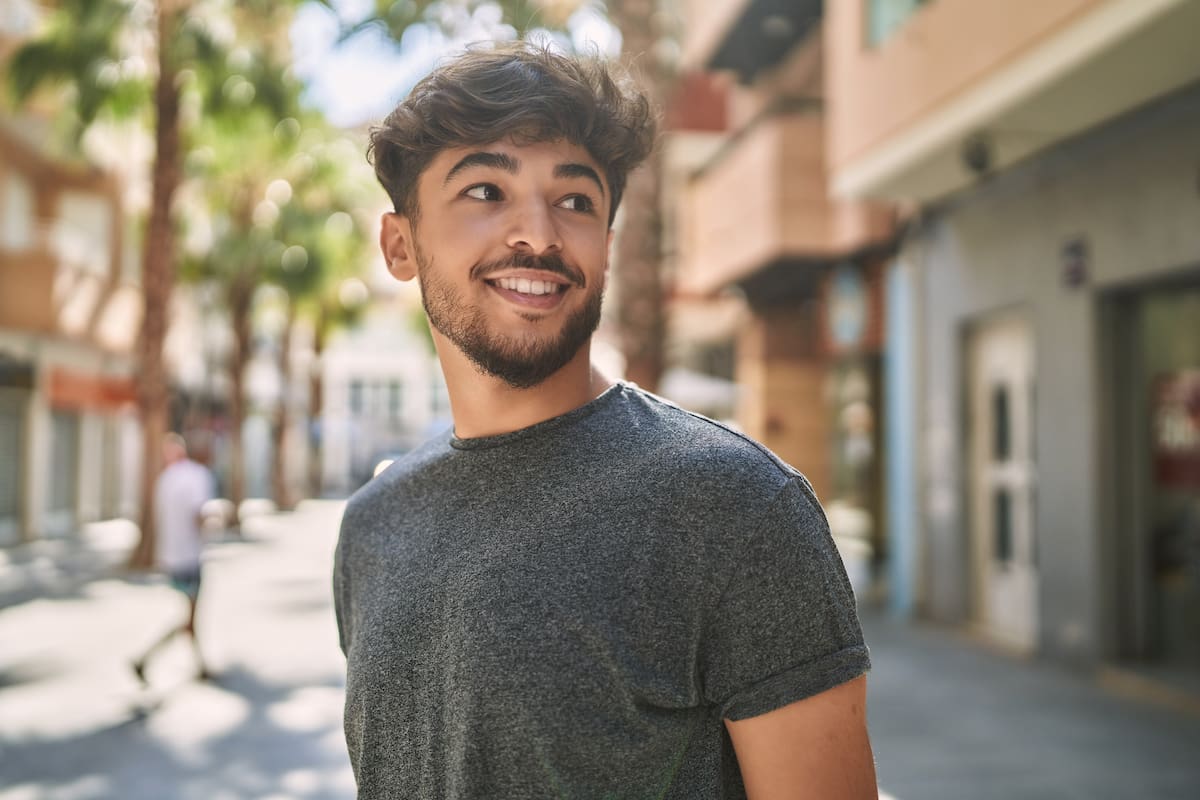 Dark haired man standing outside in the street looking to his side and smiling